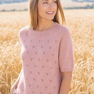 May include: A light pink short-sleeved sweater with a round neckline and a delicate openwork pattern. The sweater is made of a soft knit material. The model is wearing white pants. The background is a wheat field.