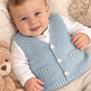 Puede incluir: Un bebé con un chaleco de punto azul claro con tres botones blancos y dos bolsillos delanteros. El bebé también lleva una camisa blanca con cuello y pantalones beige. Un osito de peluche es visible en el lado izquierdo de la imagen.