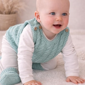 Puede incluir: Un bebé con un chaleco de punto verde azulado claro con botones de madera, una camisa blanca de manga larga y botines a juego. El chaleco tiene un diseño de punto trenzado. El bebé está gateando sobre una superficie blanca texturizada.