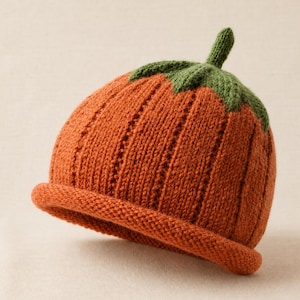 Puede incluir: Un gorro de punto en un vibrante color naranja calabaza, con una textura acanalada y un borde enrollado. La parte superior del gorro está adornada con un tallo y una hoja de punto verde, imitando la apariencia de una pequeña calabaza.