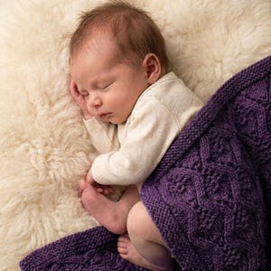 Puede incluir: Un bebé dormido con un body color crema, acurrucado sobre una superficie suave y blanquecina. Una manta de punto morado oscuro con un patrón de diamantes cubre al bebé. El bebé tiene el pelo castaño claro.