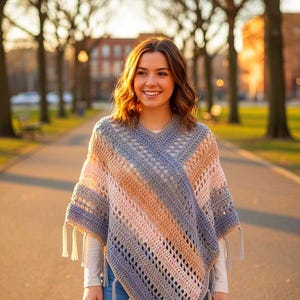 Puede incluir: Un poncho de ganchillo hecho a mano en tonos grises, rosas y beige con flecos. El poncho tiene un cuello en V y un patrón de rayas. La imagen muestra el poncho usado al aire libre en un día soleado.