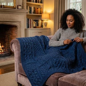 Puede incluir: Una mujer sentada en un sofá malva teje una manta azul marino de punto de cable. Una chimenea con fuego encendido está en el fondo. La habitación tiene un ambiente acogedor y acogedor, con luz natural de una gran ventana.
