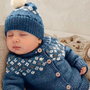Puede incluir: Un bebé dormido con un gorro de punto azul con un pompón blanco y una rebeca a juego. La rebeca tiene una parte delantera abotonada y un estampado geométrico en blanco y marrón. El bebé está acostado sobre una manta suave de color neutro.