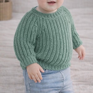 Puede incluir: Un suéter de punto verde salvia con detalles acanalados. El suéter tiene cuello redondo y mangas largas. El niño lleva vaqueros azules. El suéter es una prenda suave y cómoda.