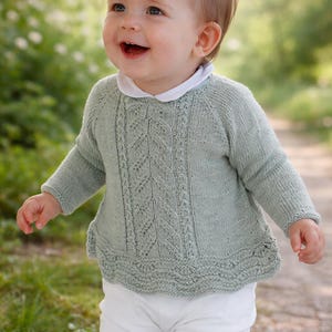 Puede incluir: Un bebé sonriente con un suéter de punto verde claro con cuello blanco y pantalones blancos. El suéter tiene un patrón de hojas detallado en la parte delantera. El bebé está afuera en un camino con follaje verde en el fondo.