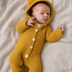 May include: A newborn baby wearing a mustard yellow knitted hooded jumpsuit. The jumpsuit has a button-up front with three white buttons and a textured knit pattern. The baby is lying on a light beige knitted blanket.