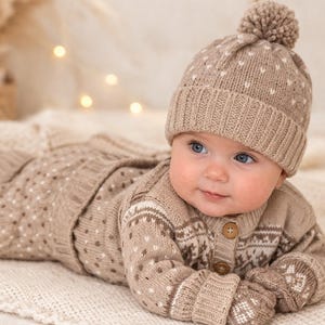 Puede incluir: Un bebé con un conjunto de punto a juego, que incluye gorro con pompón, cárdigan, pantalón y manoplas. El conjunto beige presenta un estampado de lunares blancos y diseños geométricos. El bebé está tumbado sobre una manta color crema.