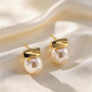 May include: A pair of gold-colored stud earrings. Each earring features a rectangular top and a large, round, cream-colored pearl. The earrings are set against a soft, off-white background.