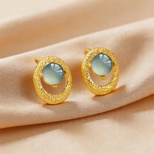 May include: A pair of gold-colored stud earrings. Each earring features a light green, round gemstone set within an oval, textured gold frame. The earrings are displayed on a beige satin surface.