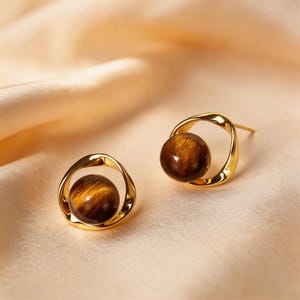 Op de afbeelding: Een paar goudkleurige oorstekers. Elke oorbel heeft een ronde, gepolijste tijgeroogsteen, gezet in een gedraaid gouden frame. De oorbellen worden gepresenteerd op een perzikroze satijnen oppervlak.