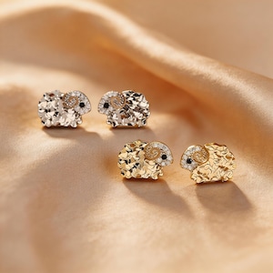 Peut inclure: Quatre paires de boucles d'oreilles en forme de bélier. Deux paires sont argentées et deux sont dorées. Chaque boucle d'oreille présente un corps texturé, une corne enroulée et un petit accent noir. Les boucles d'oreilles sont présentées sur un tissu drapé brun doré.