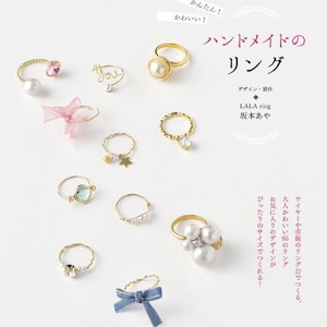 以下が含まれることがあります： 様々なデザインの手作りリングのコレクション。 金色のバンドにパール、宝石、青いリボンがあしらわれています。 リングは白い表面に展示されており、日本語のテキストとデザインの詳細が記載されています。