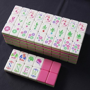 May include: A collection of white Mahjong tiles with pink and green striped edges. The tiles display colourful illustrations of flowers, plants, and Chinese characters. Some tiles have the letters "S", "N", "E", and "W" printed on them.
