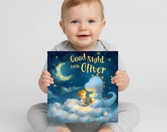 May include: A baby holding a children's book titled "Good Night little Oliver." The book cover features a blue dinosaur, a child, a crescent moon, and stars against a dark blue background. The baby is wearing a gray onesie and smiling.