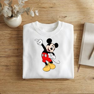 Puede incluir: Sudadera blanca de cuello redondo con un dise&ntilde;o de Mickey Mouse se&ntilde;alando. Mickey lleva pantalones cortos rojos, zapatos amarillos y guantes negros. La sudadera est&aacute; hecha de un material suave.