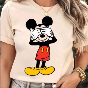 Puede incluir: Camiseta beige con una imagen de dibujos animados de Mickey Mouse cubri&eacute;ndose los ojos con manos enguantadas blancas. Mickey Mouse lleva pantalones cortos rojos con botones amarillos y zapatos amarillos. La camiseta tiene mangas cortas.