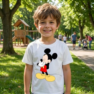 Puede incluir: Una camiseta blanca con un personaje de dibujos animados de Mickey Mouse sonriendo. El dibujo animado tiene orejas negras, una cara negra, una camisa roja, zapatos amarillos y guantes blancos. La camiseta la lleva un ni&ntilde;o en un parque.