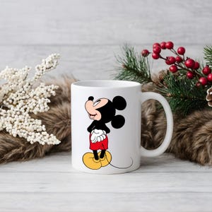 Puede incluir: Taza de cer&aacute;mica blanca con Mickey Mouse, un personaje de dibujos animados. El personaje lleva sus pantalones cortos rojos, zapatos amarillos y guantes blancos. La taza tiene un asa curvada. El fondo incluye decoraciones festivas con vegetaci&oacute;n y bayas rojas.