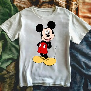 Puede incluir: Camiseta blanca con un dibujo de Mickey Mouse. El personaje de dibujos animados se muestra con orejas negras, pantalones cortos rojos, zapatos amarillos y una cara sonriente. La camiseta est&aacute; hecha de una tela suave y de color claro.
