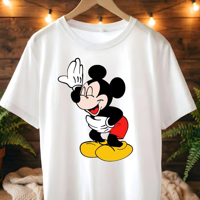 Puede incluir: Camiseta blanca con un dise&ntilde;o de Mickey Mouse sonriendo. El personaje de dibujos animados est&aacute; representado en negro, rojo y amarillo, con la mano levantada saludando. La camiseta est&aacute; colgada en una percha de madera.