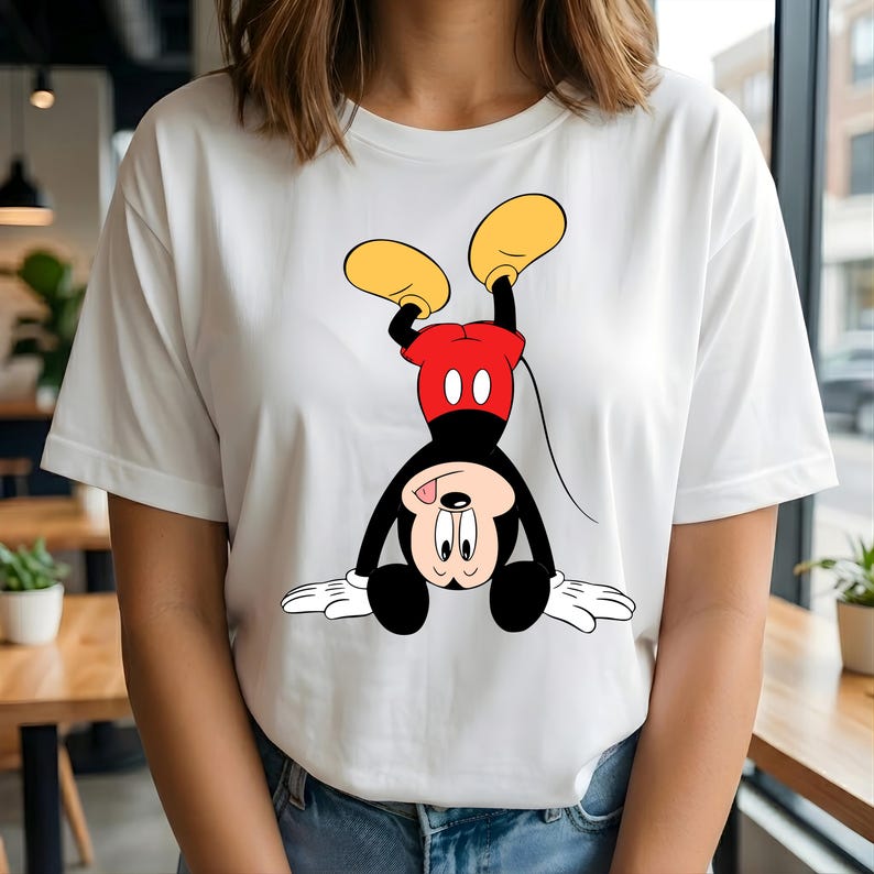 Puede incluir: Camiseta blanca con una ilustraci&oacute;n de dibujos animados de Mickey Mouse boca abajo. Mickey se representa con su cuerpo negro, pantalones cortos rojos, zapatos amarillos y guantes blancos. El dise&ntilde;o est&aacute; centrado en la camiseta.