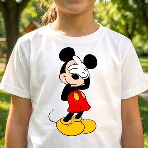 Puede incluir: Camiseta blanca con un gr&aacute;fico colorido de Mickey Mouse cubri&eacute;ndose los ojos con su mano enguantada blanca. Mickey Mouse lleva pantalones cortos rojos con botones amarillos y zapatos amarillos. El personaje de dibujos animados est&aacute; sobre un fondo verde.