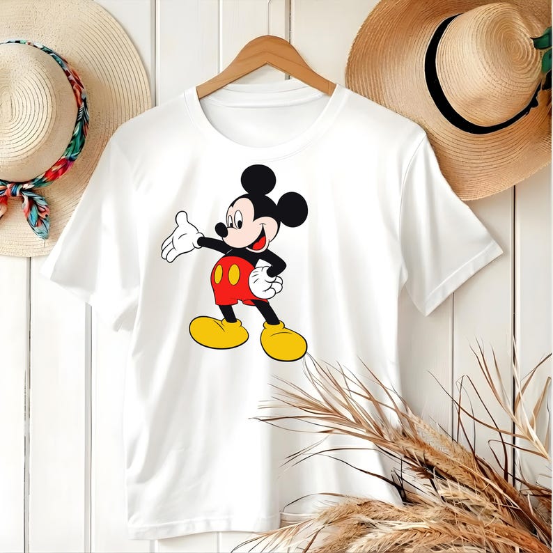 Puede incluir: Camiseta blanca con un gr&aacute;fico colorido de Mickey Mouse. El personaje de dibujos animados est&aacute; representado con su atuendo cl&aacute;sico: pantalones cortos rojos, zapatos amarillos y guantes negros. La camiseta est&aacute; colgada de una percha de madera.