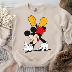 Puede incluir: Sudadera beige con un gr&aacute;fico de Mickey Mouse. El personaje de dibujos animados se muestra en una pose din&aacute;mica, con pantalones cortos rojos, zapatos amarillos y guantes blancos. La sudadera tiene cuello redondo y mangas largas.