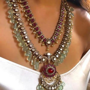 May include: Two-strand necklace set with a vintage aesthetic. The necklaces feature ruby-colored stones, small pearls, and clear, teardrop-shaped accents. The metal has a gold tone. The necklace is worn on a white top.