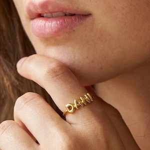 May include: A gold-toned metal ring featuring the name DANI in raised, rounded, block-style capital letters. The ring has a thin, smooth band that encircles the finger, showcasing a polished metallic finish that reflects light across the surface.