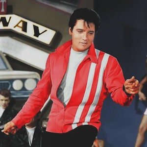 May include: A man wearing a red jacket with white vertical stripes and a white shirt underneath. The jacket has a zipper and a collar. The background includes a sign that says "WAY". The man is in a performance pose.
