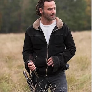 May include: Black suede jacket with a tan shearling collar, worn over a white t-shirt. The jacket has a zipper closure and is paired with dark denim jeans. The person is holding a knife and a sheath.