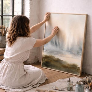 May include: A framed watercolor landscape painting with a natural wood frame. The artwork features a serene scene with muted blues, greens, and beige tones. The painting is being held up against a white wall, suggesting a gallery or studio setting.