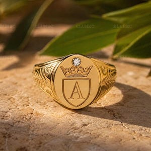 Puede incluir: Un anillo de sello de oro con una cara ovalada que presenta un escudo grabado con la letra A, rematado por un pequeño motivo de corona y una piedra preciosa transparente incrustada. La banda muestra grabados de hojas en los lados.
