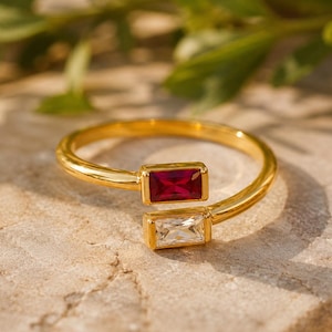 May include: A gold-toned open ring featuring two baguette-cut stones, one deep red and one clear white, set in a horizontal orientation at each end of the band. The metal band has a smooth, polished finish and an adjustable open design.