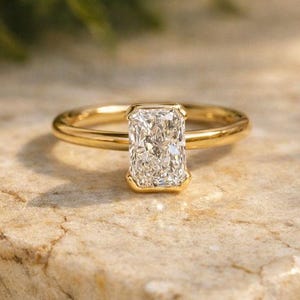 以下が含まれることがあります： 長方形のダイヤモンドが特徴のゴールドの婚約指輪。 ダイヤモンドはベゼルセッティングでセットされています。 リングは、質感のある明るい色の石の表面に表示されています。