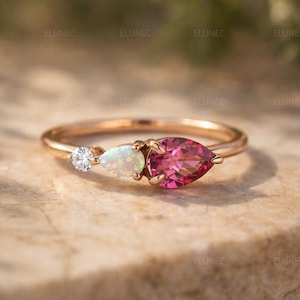 May include: A rose gold ring featuring three gemstones. A small, round, clear gemstone is on the left, followed by a pear-shaped opal, and a larger, pear-shaped, pink gemstone on the right. The ring is set on a textured surface.