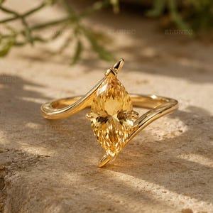 Puede incluir: Un anillo de oro con una piedra preciosa amarilla de corte marquesa montada en una banda de estilo bypass. La banda se curva alrededor de la piedra central, que muestra una superficie facetada que refleja la luz. El metal tiene un acabado pulido.