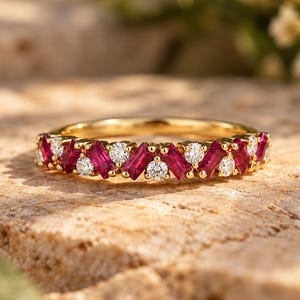 Op de afbeelding: Een gouden ring met een afwisselend patroon van rood geslepen robijnen in baguettevorm en witte ronde diamanten. De edelstenen zijn in een doorlopende rij gezet langs de bovenste helft van de gepolijste geelgouden ringband voor contrast.