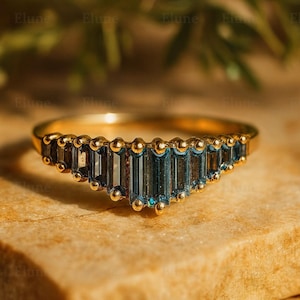 以下が含まれることがあります： 青緑色の長方形の宝石が並んだゴールドリング。リングは、各宝石の間に小さな金のビーズをあしらった繊細なデザインで、ユニークでエレガントな外観を作り出しています。リングは、質感のある明るい茶色の表面に置かれています。