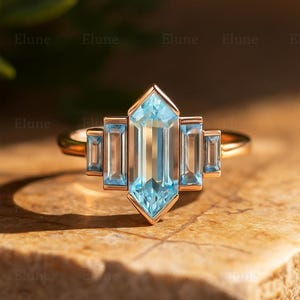 May include: A gold ring with a central, light blue, emerald-cut gemstone, and four smaller, rectangular blue gemstones. The ring has a geometric design. The word "Elune" is repeated in the background. The ring is set against a neutral background.