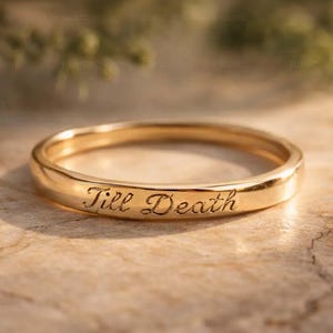May include: A gold-colored ring with the inscription "Till Death" engraved on its surface. The ring is set against a blurred background, highlighting the inscription. The ring is a symbol of commitment.