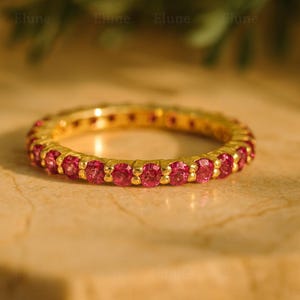 Puede incluir: Un anillo de la eternidad dorado con una fila continua de gemas rosas vibrantes. El anillo se coloca sobre un fondo neutro, resaltando el brillo de las gemas. El anillo es una pieza de joyería.