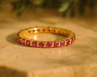 Könnte beinhalten: Ein goldfarbener Eternity-Ring mit einer durchgehenden Reihe leuchtend rosa Edelsteine. Der Ring ist vor einem neutralen Hintergrund platziert, der das Funkeln der Edelsteine hervorhebt. Der Ring ist ein Schmuckstück.