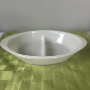 May include: A white, oval-shaped divided serving dish. The dish is made of a glossy material and has a raised rim. The interior is split into two sections by a central divider. The dish sits on a green, textured surface.