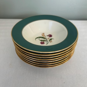 May include: A stack of seven round ceramic soup bowls featuring a wide dark green rim and a gold-trimmed edge. The center of each bowl is off-white and decorated with a small, delicate floral motif in shades of purple, green, and yellow.