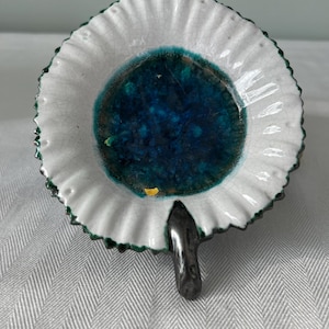 May include: A ceramic dish with a white, ruffled edge and a deep teal center. The dish has a dark gray, leaf-shaped handle. The rim is trimmed with green. The dish has a crackled glaze.