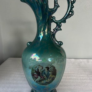 May include: A teal iridescent ceramic pitcher with a ruffled rim and an ornate, pierced handle. The body features a central circular decal depicting two children sitting outdoors with a small dog. The finish has a metallic, multi-colored sheen.