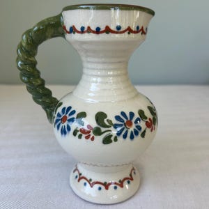 May include: A cream-colored ceramic pitcher with a crackled glaze finish, featuring a twisted green handle. The body is decorated with hand-painted blue flowers, green leaves, and red accents, with a scalloped red and blue line pattern at the rim and base.